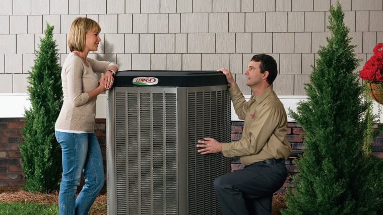 A woman talks to a technician kneeling next to a large outdoor HVAC unit between two small evergreen trees beside a house.