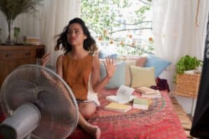Woman sits cross-legged on a bed in front of a fan, tossing her hair, surrounded by open books and colorful pillows in a bright, cozy room.
