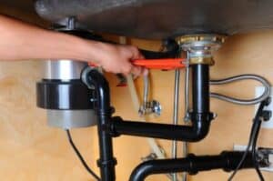 A person uses a wrench to tighten a pipe connection under a sink, with visible plumbing lines and a garbage disposal unit attached.