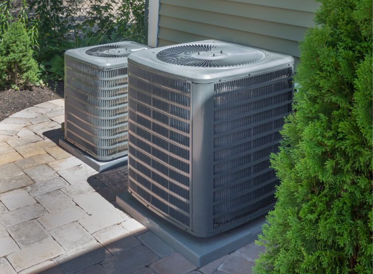 Two outdoor air conditioning units are placed on concrete pads next to a house with siding and surrounded by landscaping.