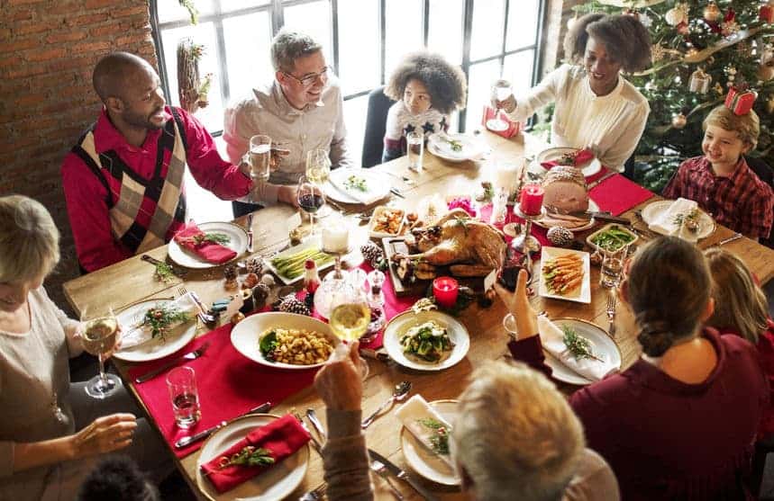 A group of people sit around a festive table with holiday decorations, enjoying a meal together near a Christmas tree.