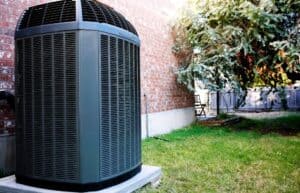 Large outdoor air conditioning unit installed next to a brick house, with green grass and a tree in the background.