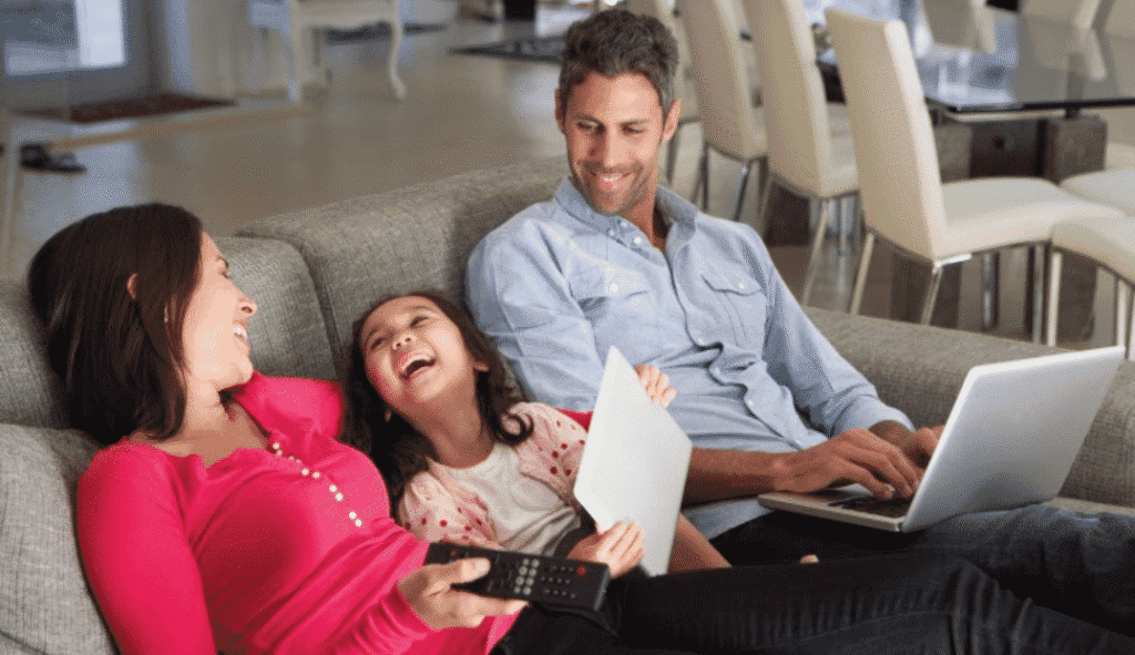A woman, a child, and a man sit on a couch. The woman holds a TV remote, the child laughs holding paper, and the man uses a laptop. Dining chairs and a table are visible in the background.