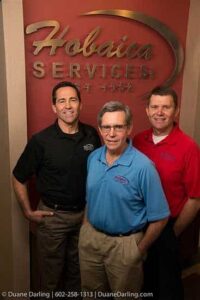 Three men standing in front of a "Hobaica Services" sign, wearing branded shirts in black, blue, and red, posing and smiling for the camera.