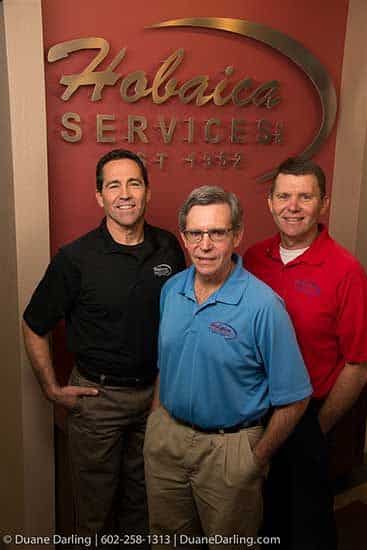Three men standing in front of a "Hobaica Services" sign, wearing branded shirts in black, blue, and red, posing and smiling for the camera.