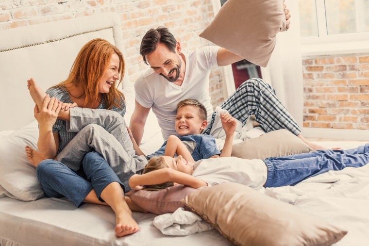 A family of four in pajamas smiles and plays with pillows on a bed in a bright bedroom.