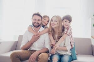 A man and woman sit on a couch, smiling, with a young girl and boy hugging them from behind.