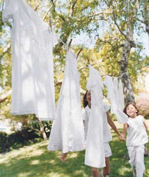 Two children stand by a clothesline with white garments hanging to dry outside on a sunny day, with trees and grass in the background.