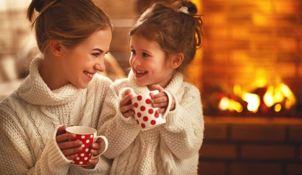 Woman and child in white sweaters smile at each other, holding red polka dot mugs, with a lit fireplace in the background.