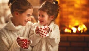 Woman and child in white sweaters smile at each other, holding red polka dot mugs, with a lit fireplace in the background.