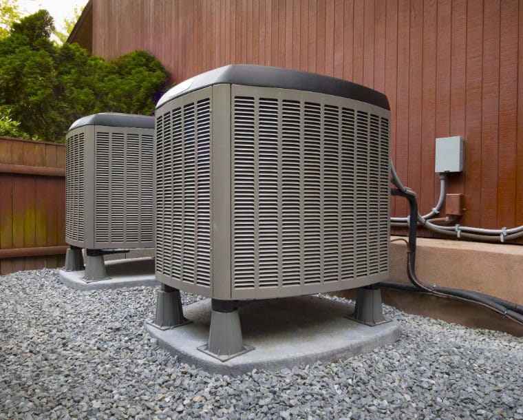 Two outdoor air conditioning units sit on cement pads next to a wood-paneled building; gravel and wiring are visible around the units.