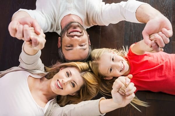 A man, woman, and young girl lie on a wooden floor, holding hands and smiling while looking up at the camera.
