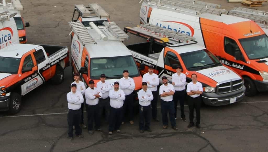A group of uniformed workers stands in front of several orange and white work trucks in a parking lot.