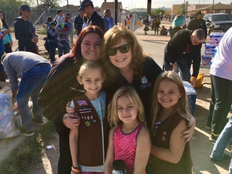 Two adults and three children pose and smile at an outdoor event with several people and tables in the background.