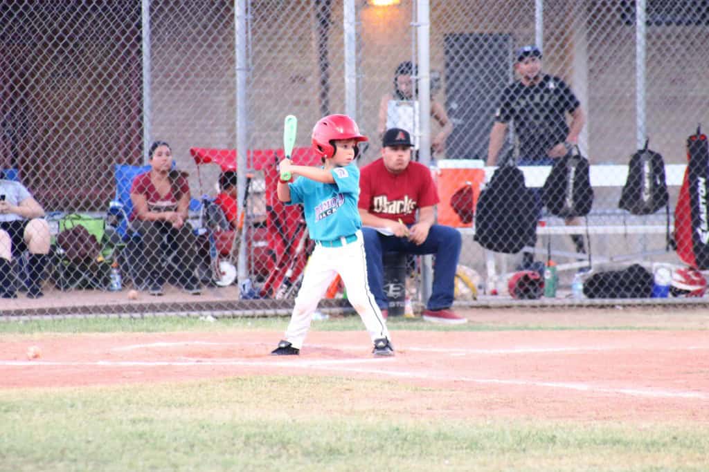 A young boy in a turquoise jersey and red helmet stands ready to bat at a baseball game while spectators watch from behind a chain-link fence.