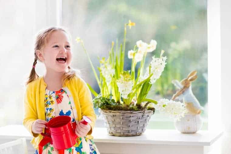 Young girl in a floral dress and yellow cardigan holds a red watering can, smiling beside a basket of blooming white flowers on a windowsill.