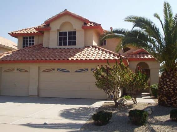 Two-story house with a red tile roof, beige exterior, three-car garage, palm tree, and desert landscaping in front.