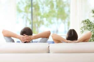 Two people sit on a white couch with their hands behind their heads, facing away from the camera, looking out a large window with greenery visible outside.