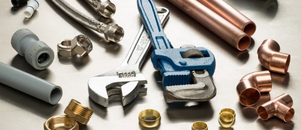 Assorted plumbing tools and fittings, including a wrench, pipe cutter, metal pipes, hoses, and various connectors, arranged on a metallic surface.