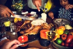 People are carving a roasted turkey at a dinner table filled with food, candles, drinks, and vegetables, with several hands and a child visible in the scene.
