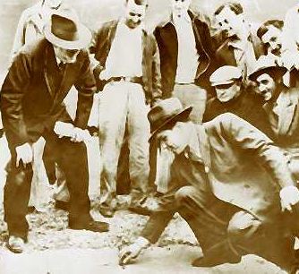 A group of men in suits and hats gather around as two men crouch on the ground, appearing to examine or play something.