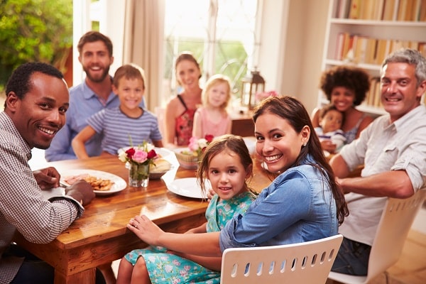 A group of adults and children sit around a dining table sharing a meal, smiling at the camera in a bright, homey room with shelves and windows in the background.