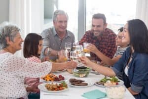 Six people of varying ages sit around a dining table, clinking glasses and smiling, with plates of food and salad bowls in front of them.