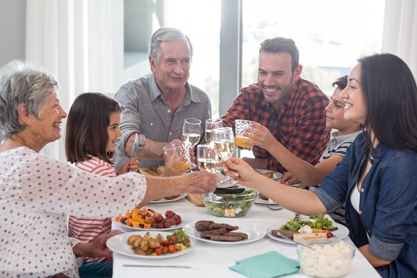 Six people of varying ages sit around a dining table, clinking glasses and smiling, with plates of food and salad bowls in front of them.