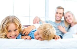 Two young children lie on a bed in the foreground, smiling, with two adults sitting in the background, all appearing relaxed in a bright room.