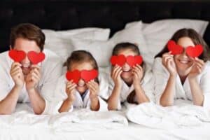 A family of four lies on a bed, each holding a red paper heart in front of their face, wearing white robes and smiling.
