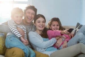 Family of four sitting on a couch, smiling and watching TV together; the girl holds the remote control.