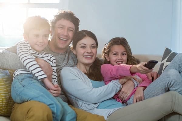 Family of four sitting on a couch, smiling and watching TV together; the girl holds the remote control.