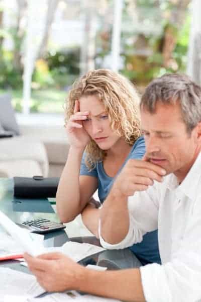 A man and woman sit at a table looking at papers with concerned expressions, a calculator and documents are spread out in front of them.