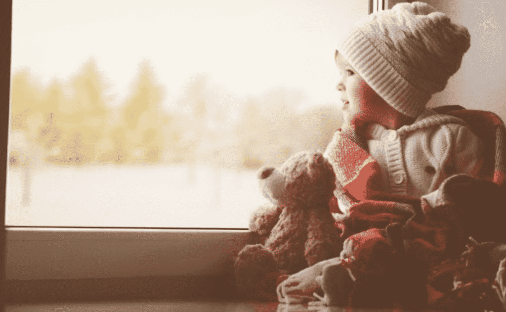 A young child in a knit hat and sweater sits by a window with a teddy bear, looking outside with a blanket around them.
