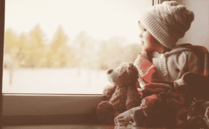 A young child in a knit hat and sweater sits by a window with a teddy bear, looking outside with a blanket around them.