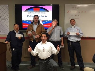 Five men stand indoors, some holding checks and cutout faces. A screen behind them reads “CONGRATS YOU WON! $30 Honda Bucks.” A whiteboard is on the right side of the image.