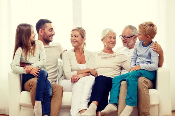 Three adults, two seniors, and two children sit together on a white couch, smiling and interacting with each other in a brightly lit room.