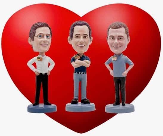 Three male bobblehead figurines stand in front of a large red heart, each holding a small American flag.