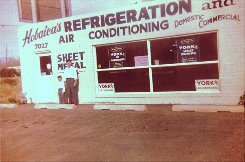 A man and a child stand outside a building with signage reading “Hobaica’s Refrigeration and Air Conditioning” and “York” in large letters.