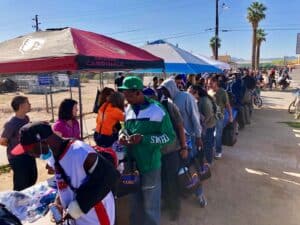 People stand in a long line outdoors under tents, waiting to receive clothing and supplies from volunteers at a distribution event.