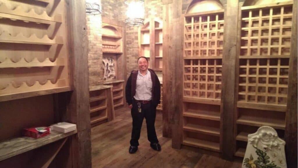 A person stands in a room with wooden wine racks, shelves, and a rustic brick wall. Warm ceiling lights illuminate the space.