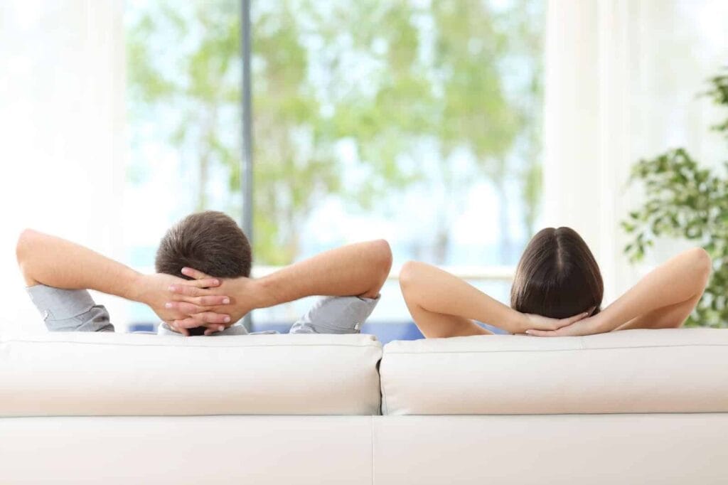 Two people sit on a white couch with their backs to the camera, arms resting behind their heads, looking out a bright window with greenery outside.
