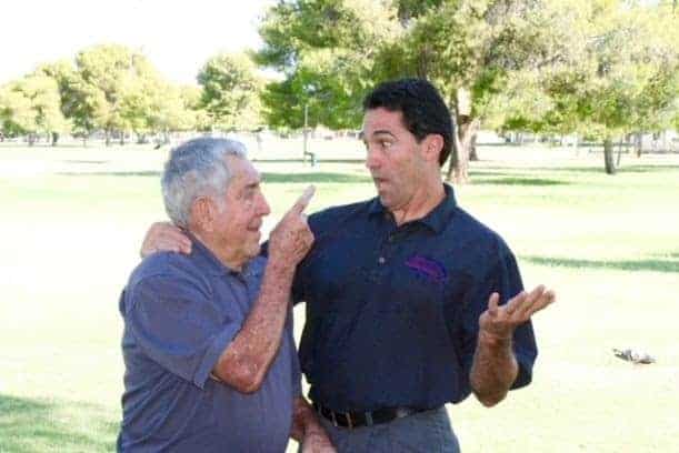 Two men stand outdoors on grass; the older man points at the younger man's face while the younger man looks surprised and gestures with one hand. Trees are visible in the background.