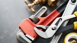 A red pipe wrench, adjustable pliers, brass valve, measuring tape, and plumbing tape arranged on a gray surface.