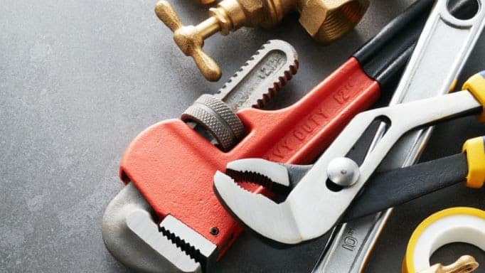 A red pipe wrench, adjustable pliers, brass valve, measuring tape, and plumbing tape arranged on a gray surface.