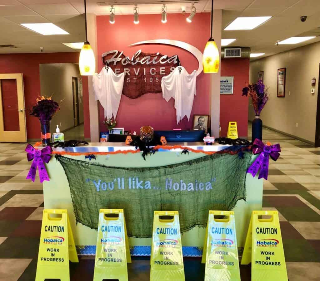 Reception desk decorated for Halloween with ghosts, pumpkins, and caution signs reading “Hobaica Work in Progress” on the floor in front of the counter.