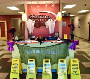 Reception desk decorated for Halloween with ghosts, pumpkins, and caution signs reading “Hobaica Work in Progress” on the floor in front of the counter.