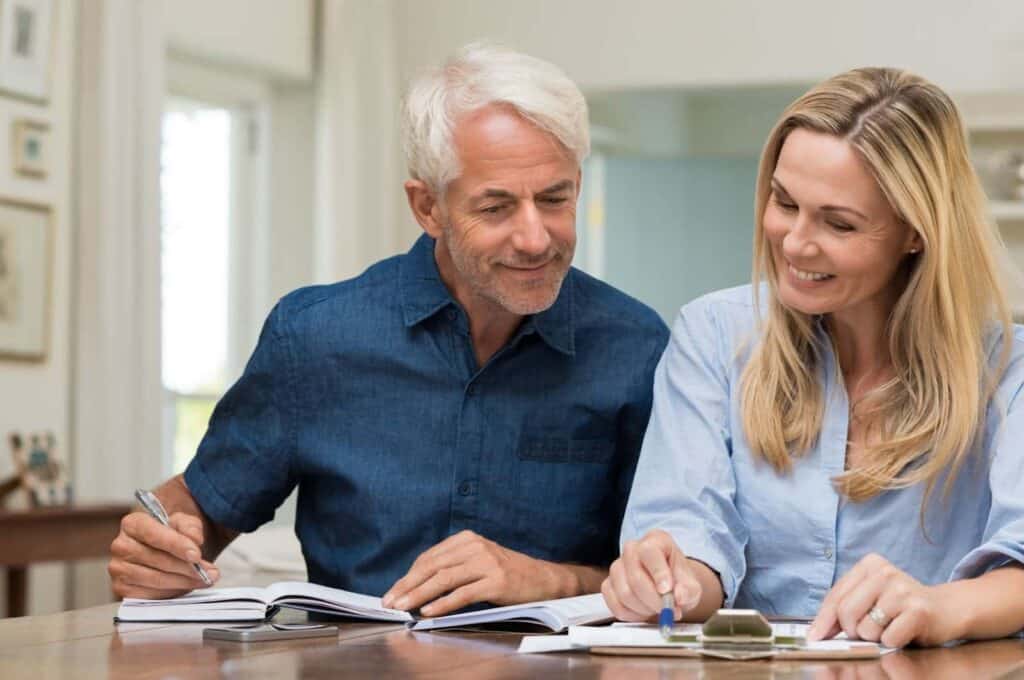 An older man and a woman sit at a table, reviewing documents together and smiling, with pens and notebooks in front of them.