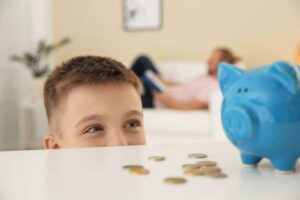 A young boy looks at a blue piggy bank and scattered coins on a table, while an adult relaxes on a couch in the background.