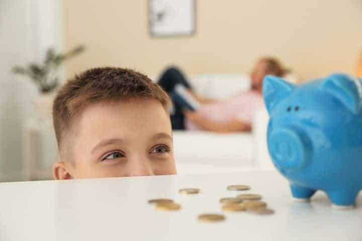 A young boy looks at a blue piggy bank and scattered coins on a table, while an adult relaxes on a couch in the background.